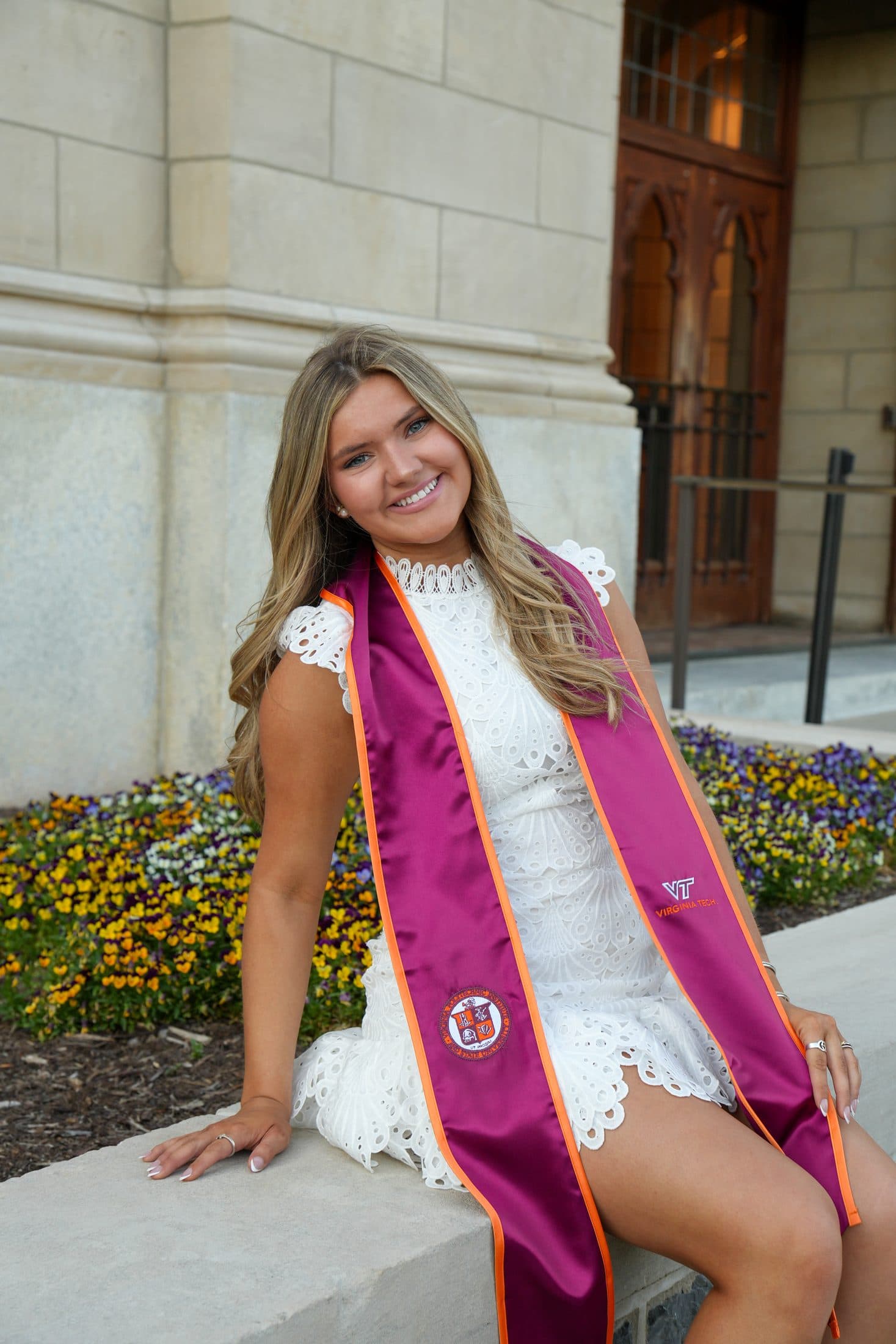 A graduate in a white lace dress framed by a wall of blooming flowers