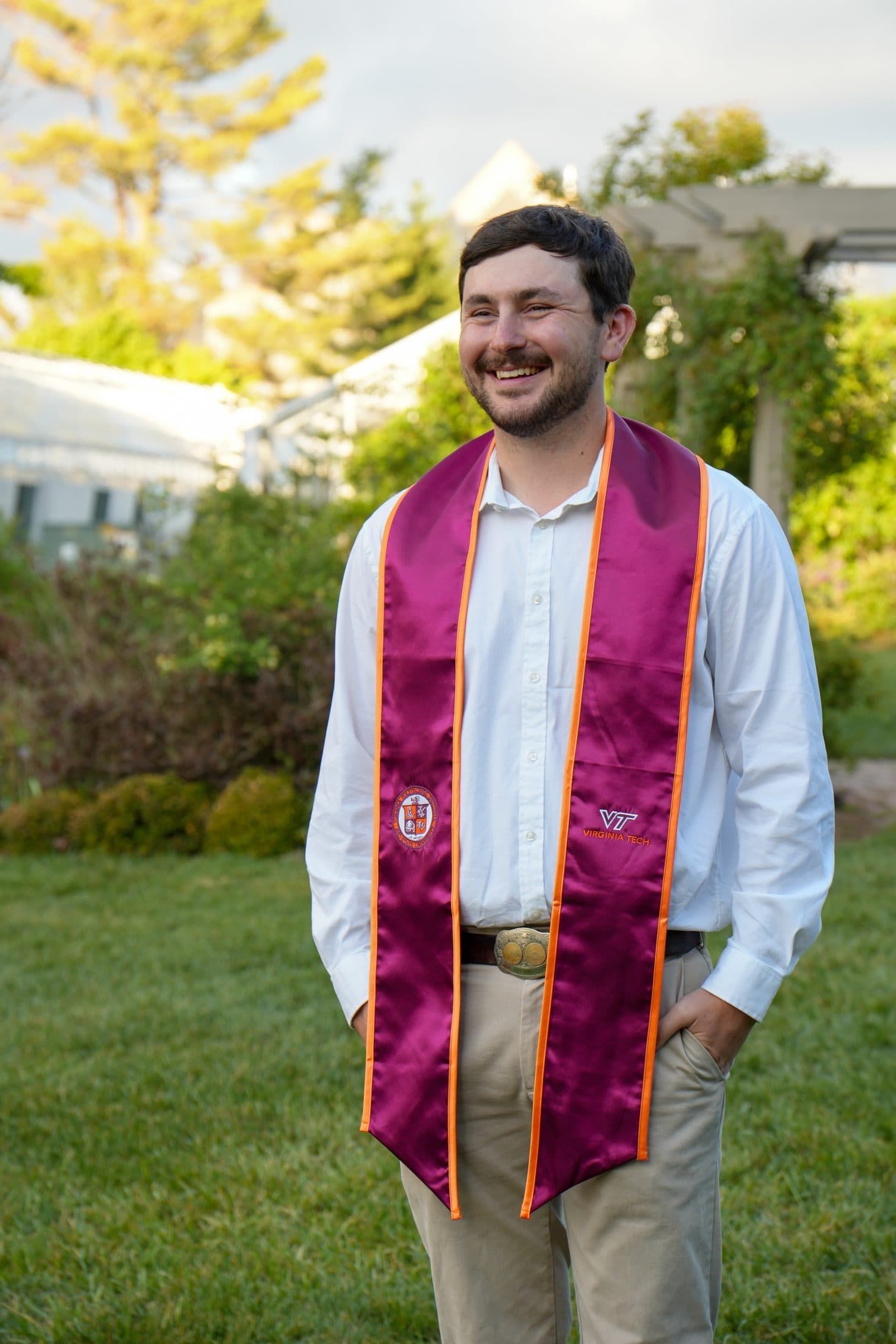 A senior in his maroon stole laughing in a green garden at dusk